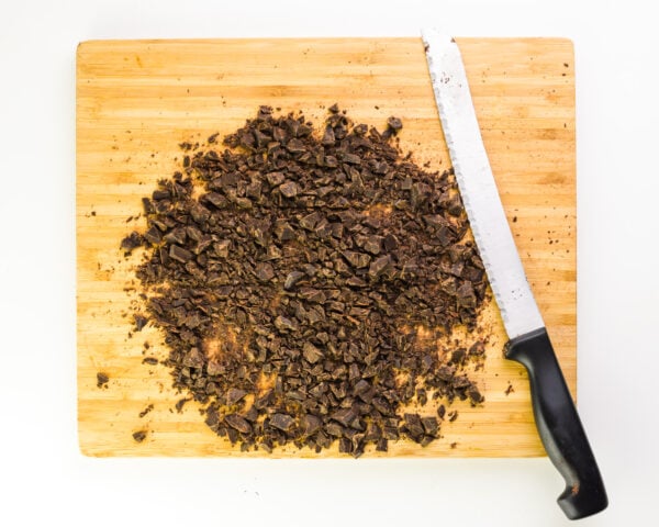 Chopped dark chocolate pieces scattered on a wooden cutting board next to a serrated knife.