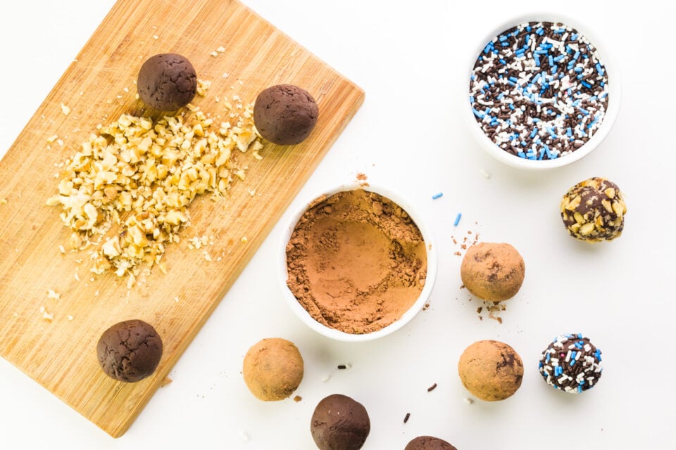 Dark chocolate balls sit next to bowls of cocoa powder and sprinkles. A cutting board has chopped nuts on it.