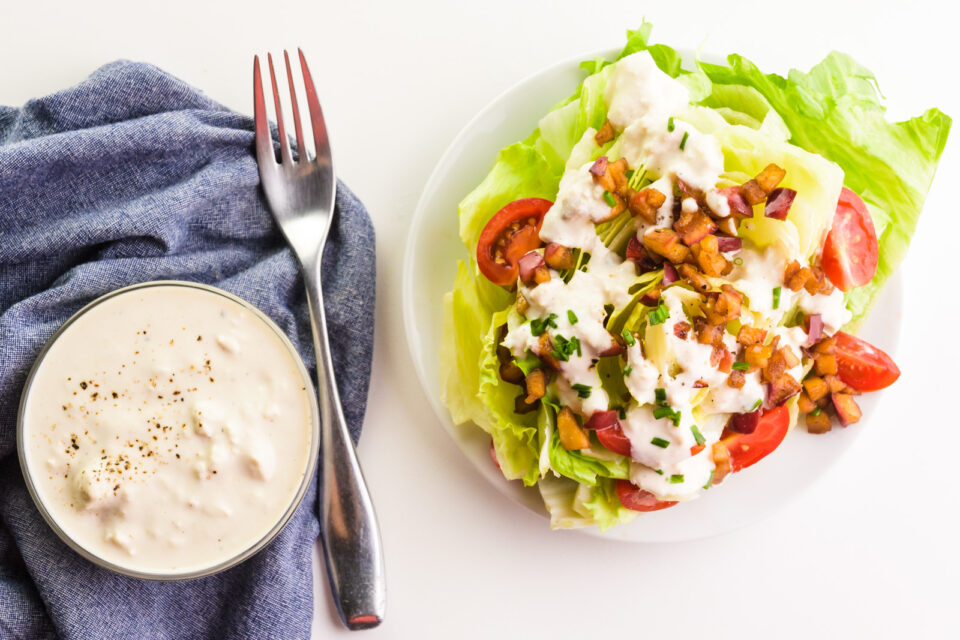 Looking down on a salad with a fork and a bowl of dressing beside it on a dark blue cloth napkin.