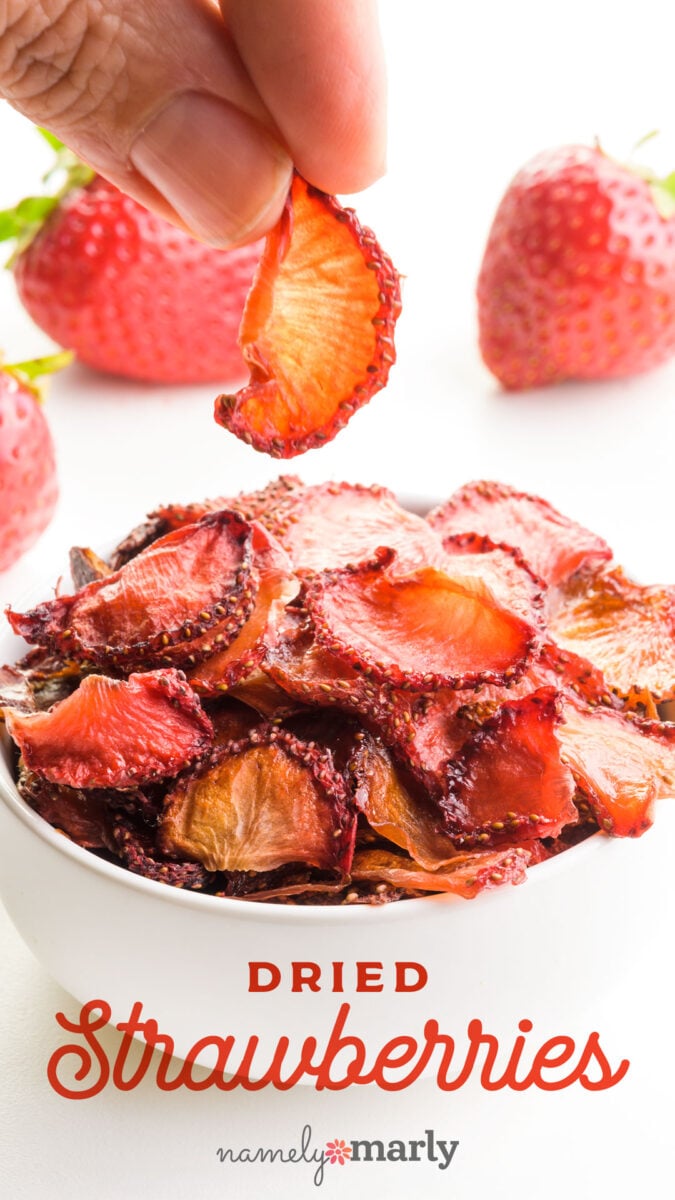 Bowl of dried strawberries with a hand holding one slice, fresh strawberries in the background.