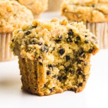 An elderberry muffin has a bite taken out, showing lots of berries. There are more muffins behind it.