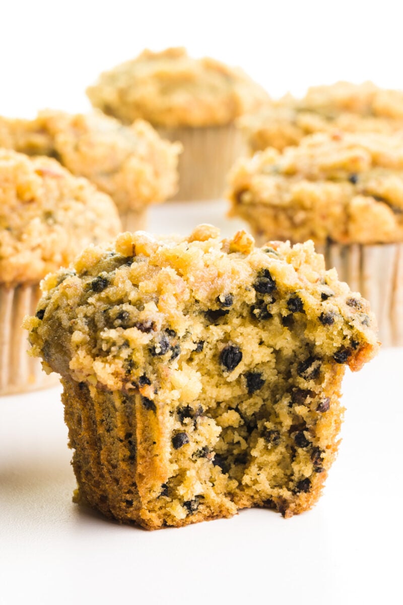 An elderberry muffin with a bite taken out sits on a white table top. There are several more muffins behind it.