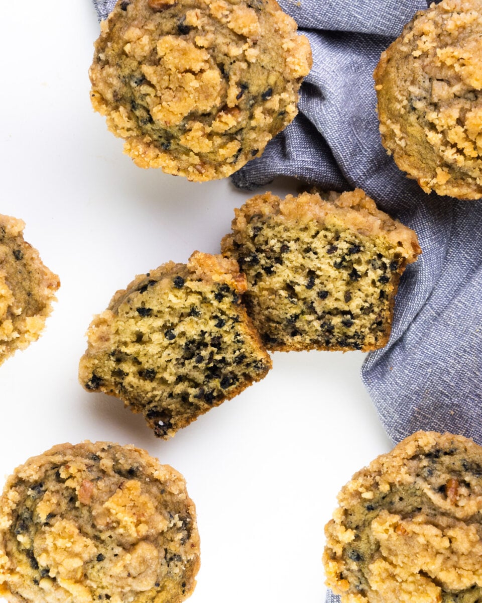 Looking down on a white table with several elderberry muffins. The one in the middle is cut in half. There's a grey kitchen towel on the right.