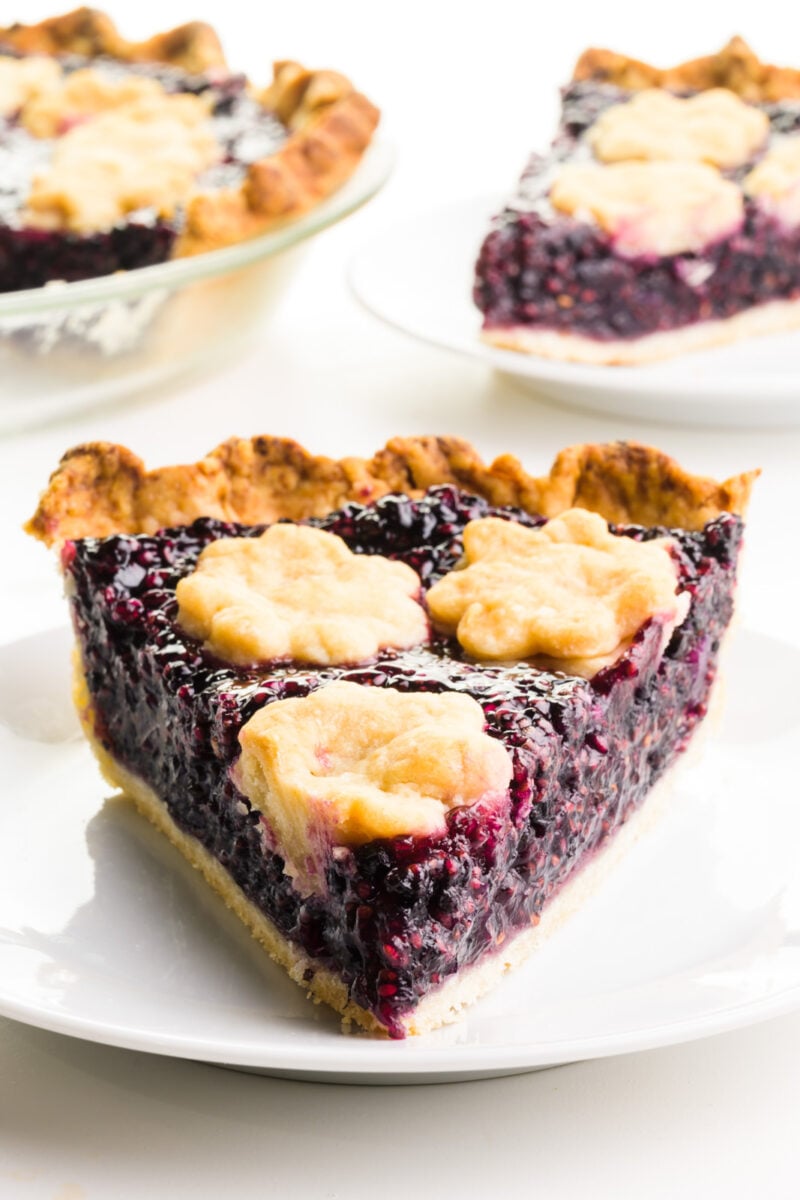 A slice of elderberry pie on a plate sits in front of another slice and the rest of the pie in the background.