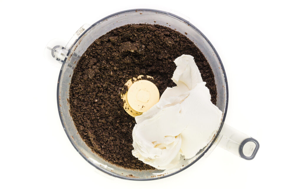 Vegan cream cheese has been added to a food processor bowl with chocolate cookie crumbs.
