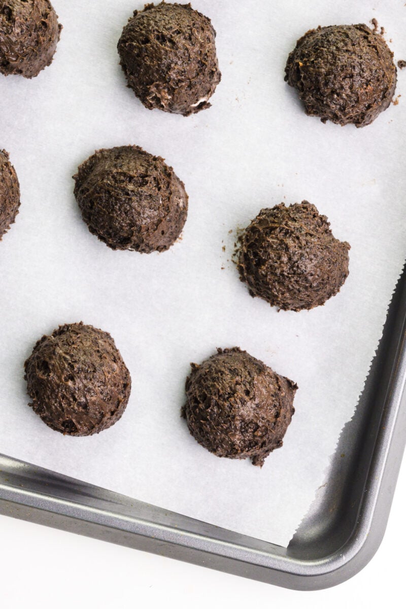 Balls of chocolate dough are lined up on a baking sheet lined with parchment paper.