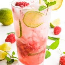 A glass holds a red beverage with slices of lime, mint, and raspberries both in the glass and around the glass too.