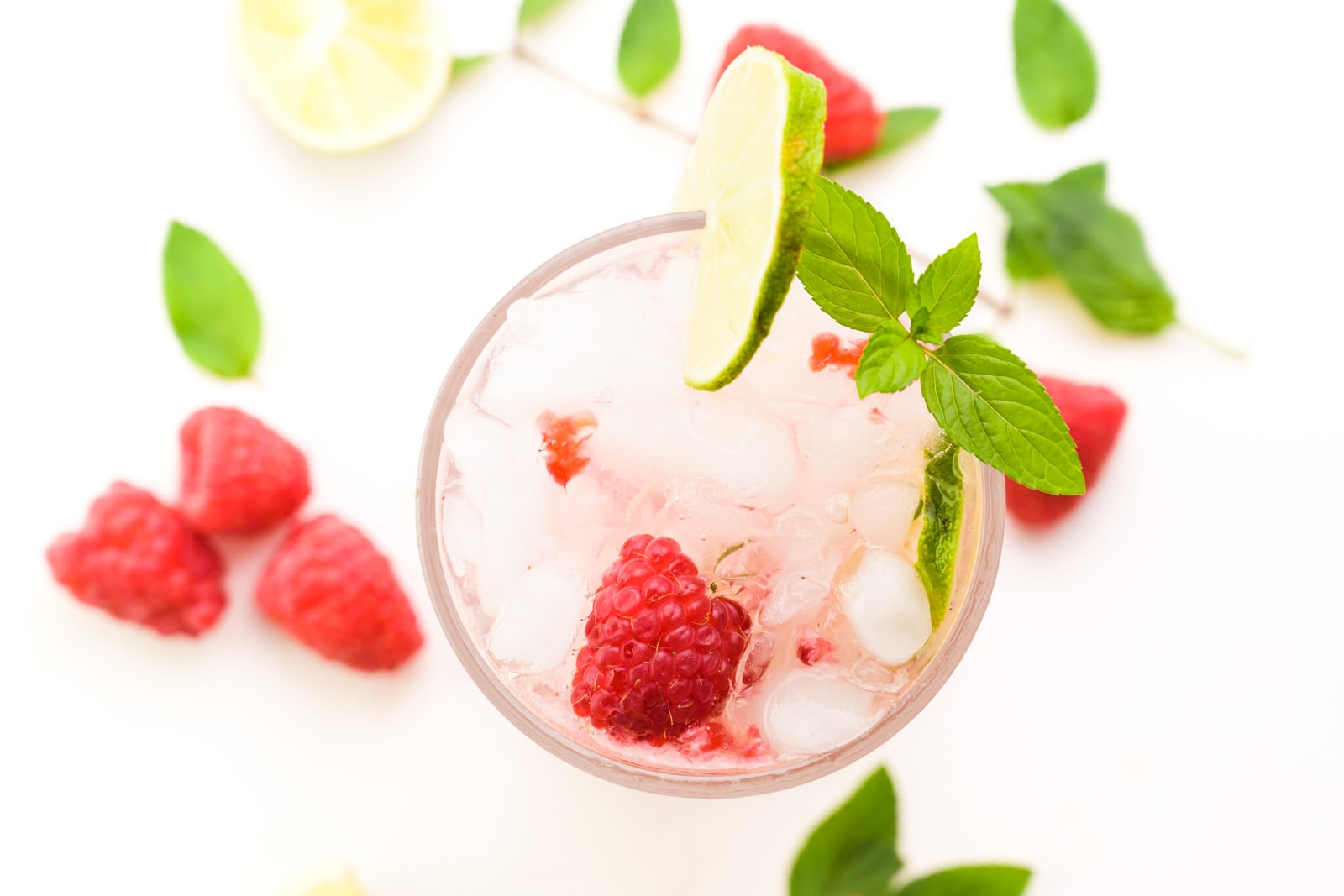 Looking down on a glass with a pink beverage inside. There's fresh raspberries, limes, and mint sprigs in the glass and around it.