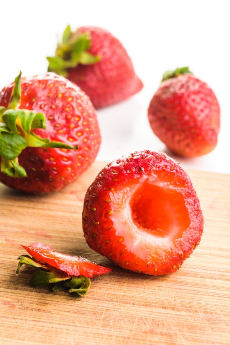 A strawberry has had the stem chopped off and the center carved out. It's sitting on a cutting board with other whole strawberries.