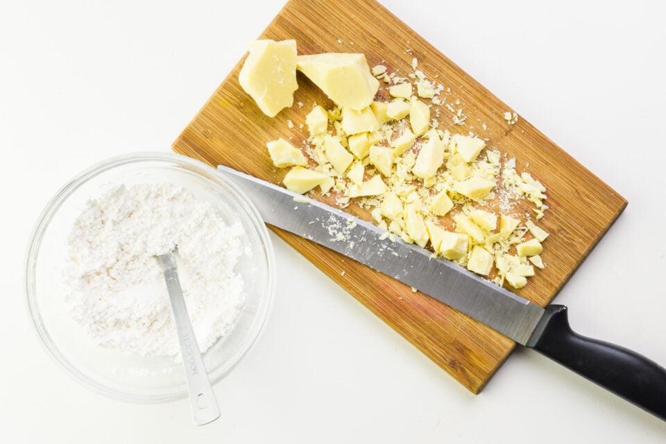 Looking down on a cutting board with a knife and chopped cocoa butter chunks. There's a bowl next to it with powdered sugar and a measuring spoon.
