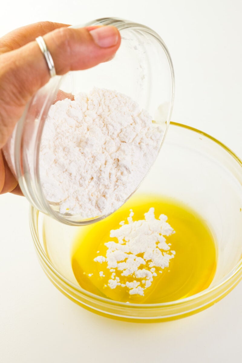 A hand holds a glass bowl of powdered sugar and is pouring it into a bowl with melted cocoa butter.
