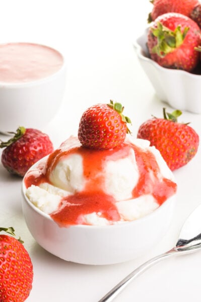 A bowl of ice cream has strawberry puree on top with fresh strawberries. There are bowls of strawberries and more sauce behind it.