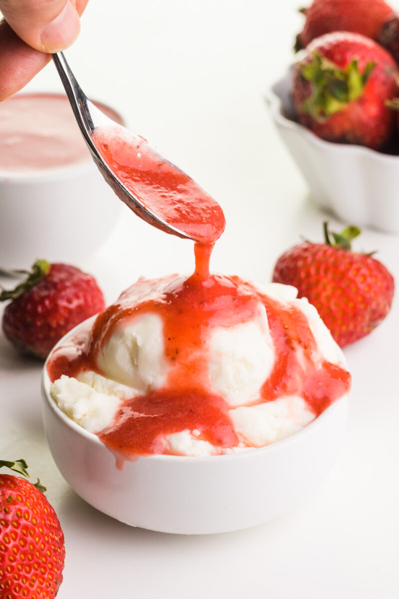 A hand holds a spoon drizzleling strawberry sauce over ice cream. There are fresh strawberries and a bowl of more sauce in the background.
