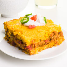 A slice of cornbread casserole sits on a plate. It's topped with sour cream and sliced tomatoes and jalapeños.