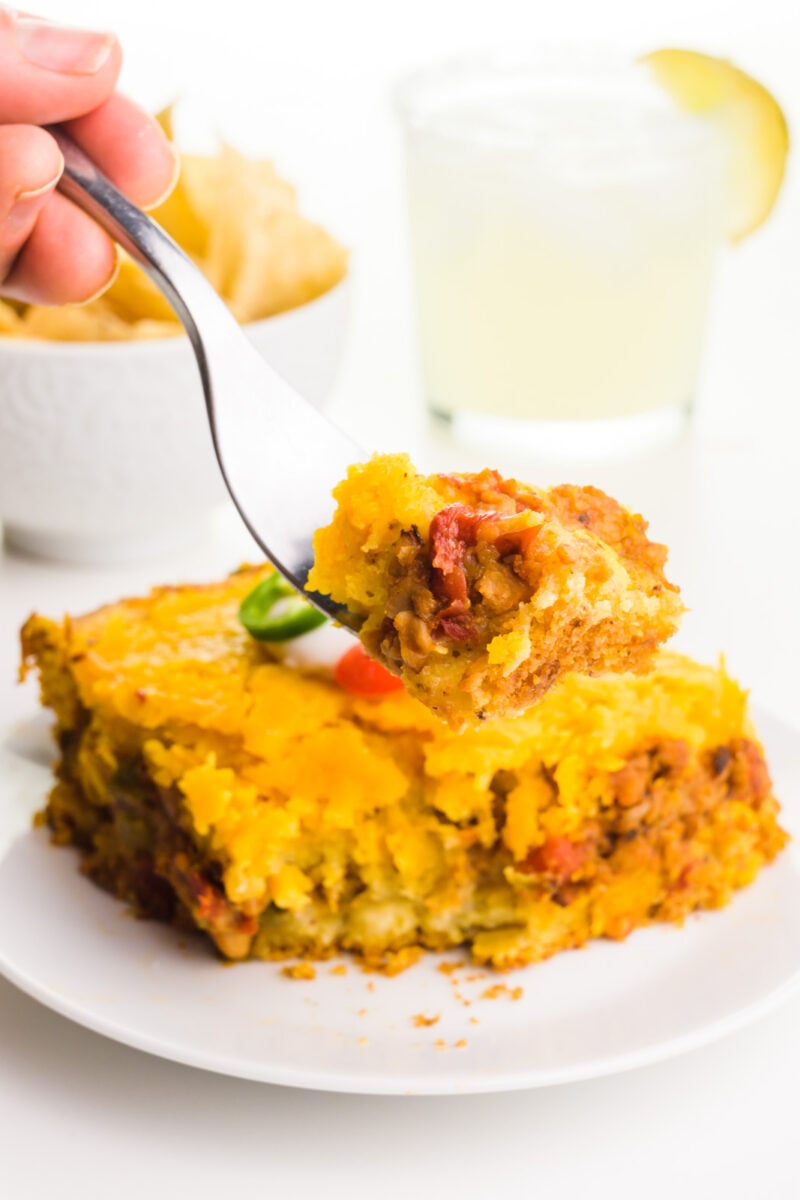 A hand holds bite of casserole, hovering over the rest of the slice. There's a bowl to tortilla chips and a margarita in the background.