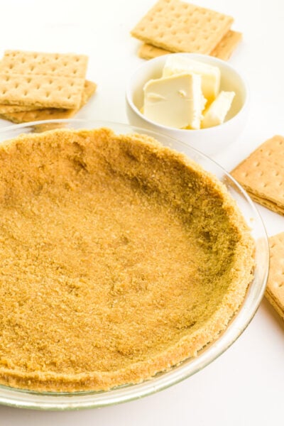 A graham cracker crust is in a glass pie pan. There are graham crackers and vegan butter around it.
