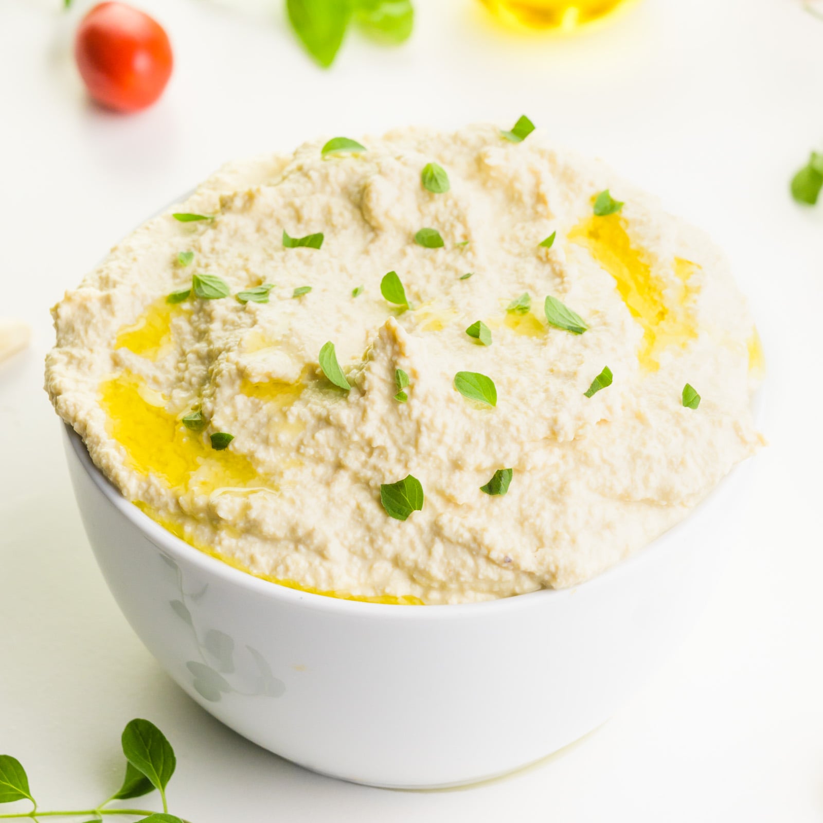 A bowl of vegan ricotta has fresh herbs on top. there are tomatoes and herbs around it.