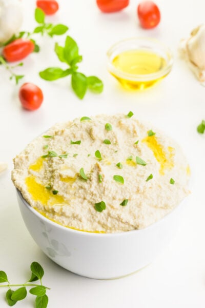 A bowl holds vegan ricotta with fresh herbs on top. There are cherry tomatoes and herbs behind it, along with a bowl of olive oil.