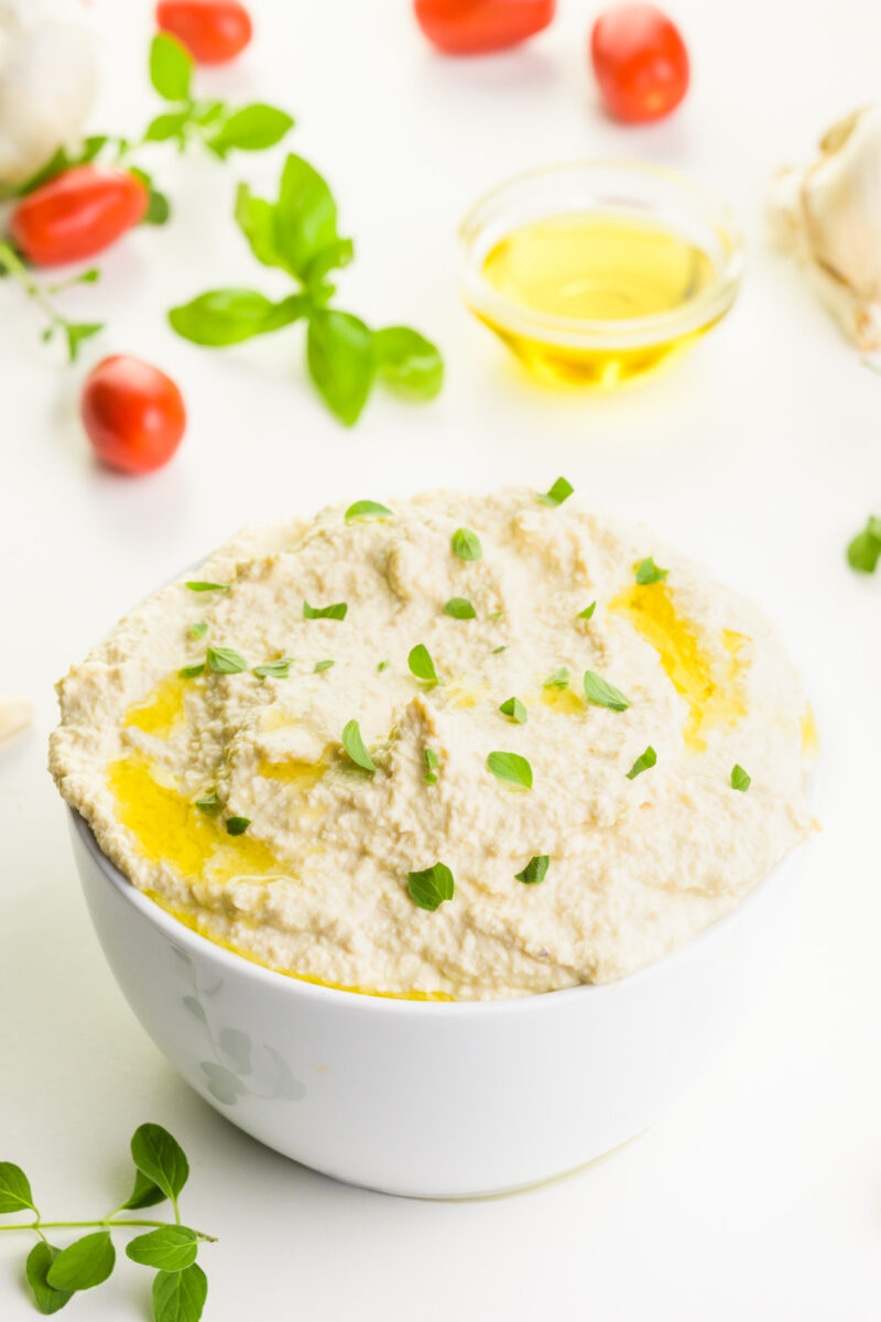 A bowl holds vegan ricotta with fresh herbs on top. There are cherry tomatoes and herbs behind it, along with a bowl of olive oil.