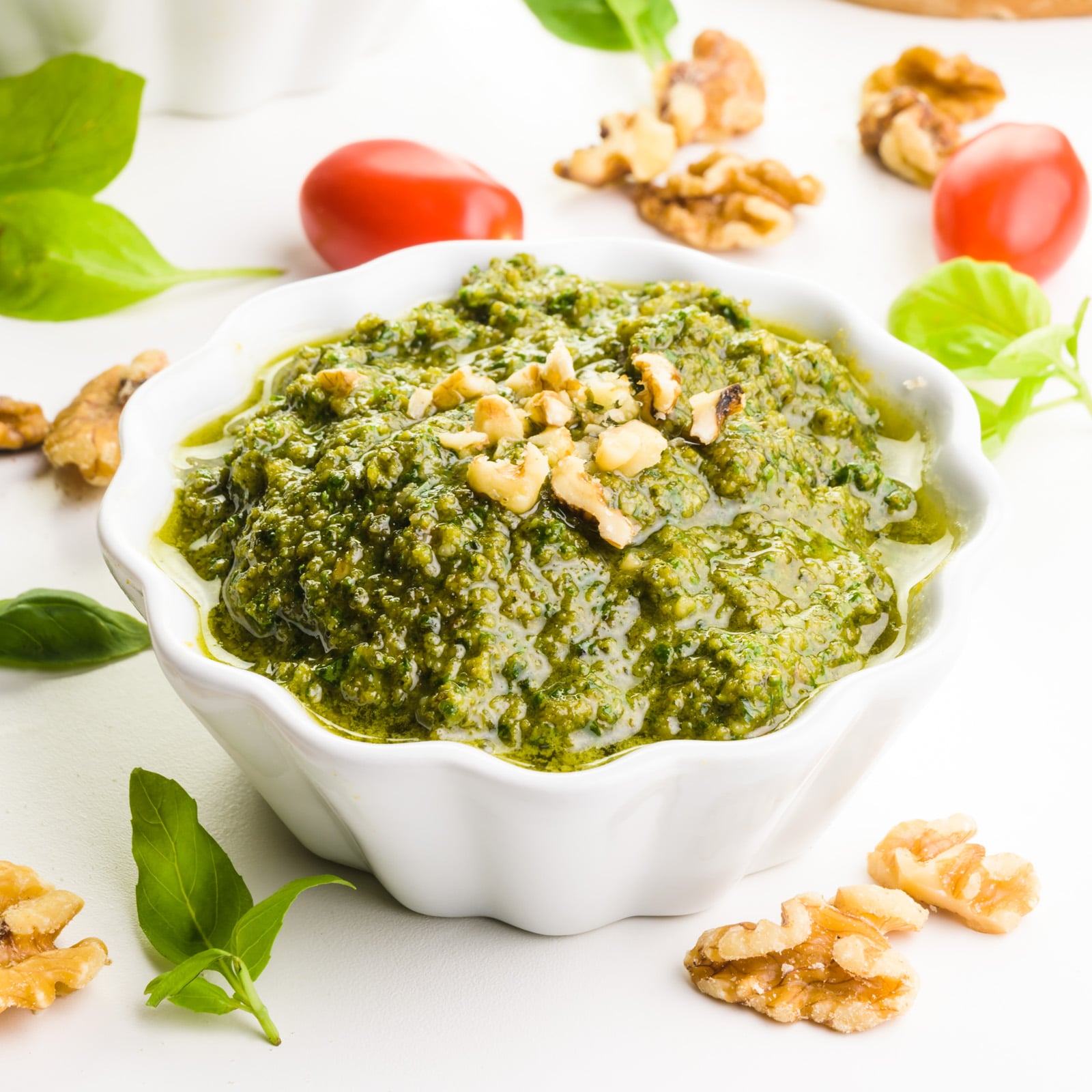 A bowl of walnut pesto has chopped walnuts on top. It's surrounded by walnuts, fresh basil leaves, and cherry tomatoes.