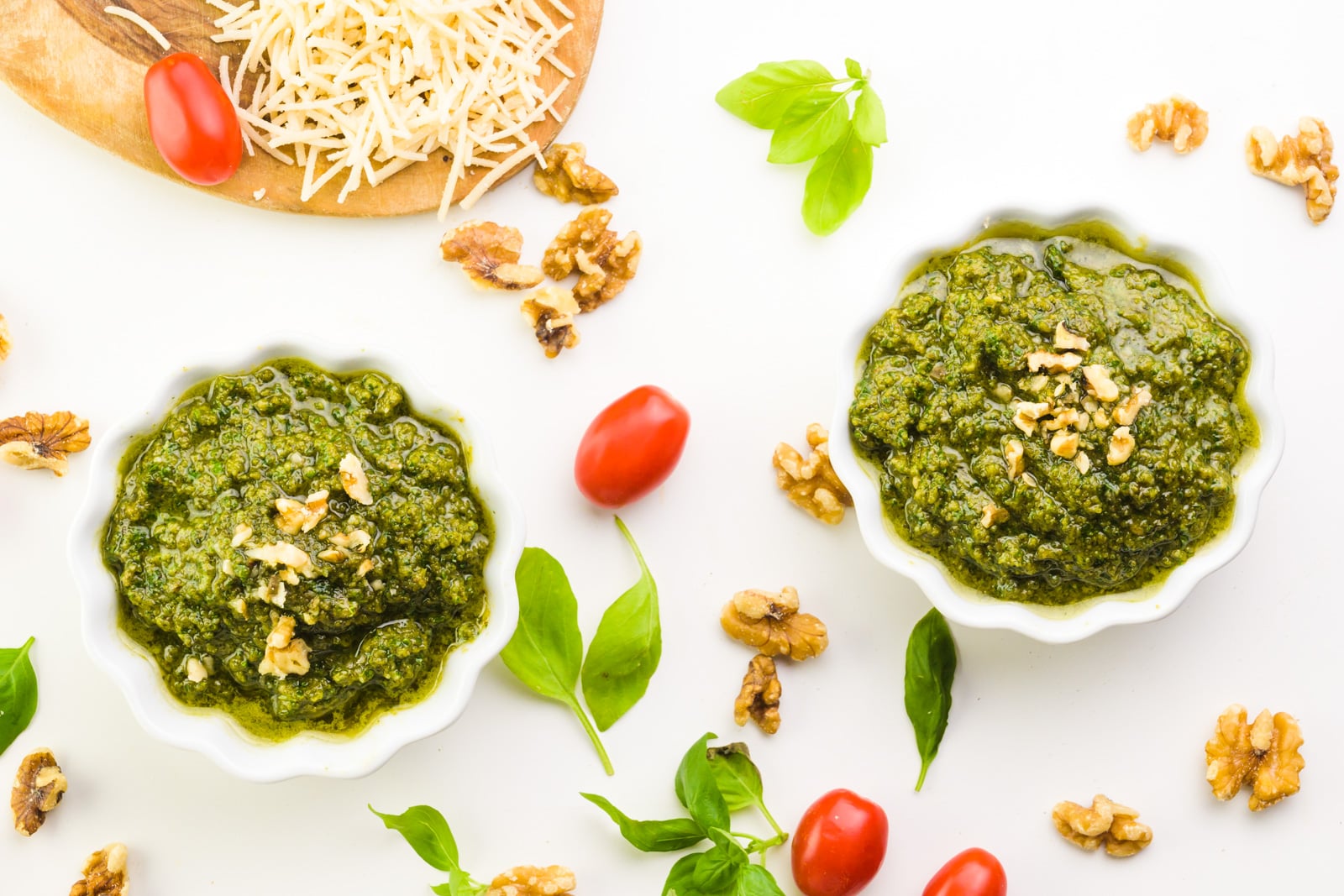 Looking down on two bowls of green sauce (pesto). There are cherry tomatoes and fresh basil leaves, along with walnuts around it.