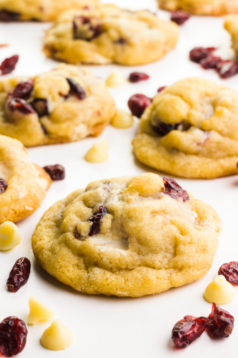 Several chocolate chip cranberry cookies sit next to each other. There are dried cranberries and white chocolate chips around them.