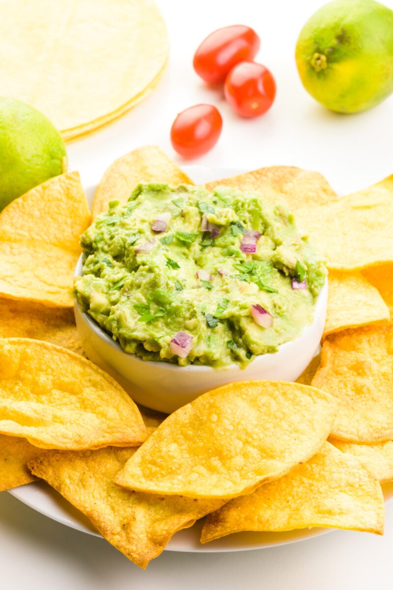 Air fryer tortilla chips are on a plate surrounding a bowl of guacamole. Behind it are fresh lemons, cherry tomatoes, and more tortillas.