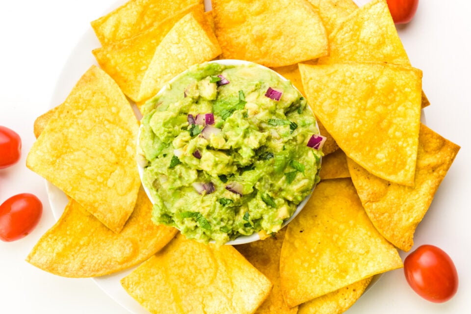 Looking down on a bowl of guacamole surrounded by air fried tortilla chips. There are cherry tomatoes and fresh limes nearby.
