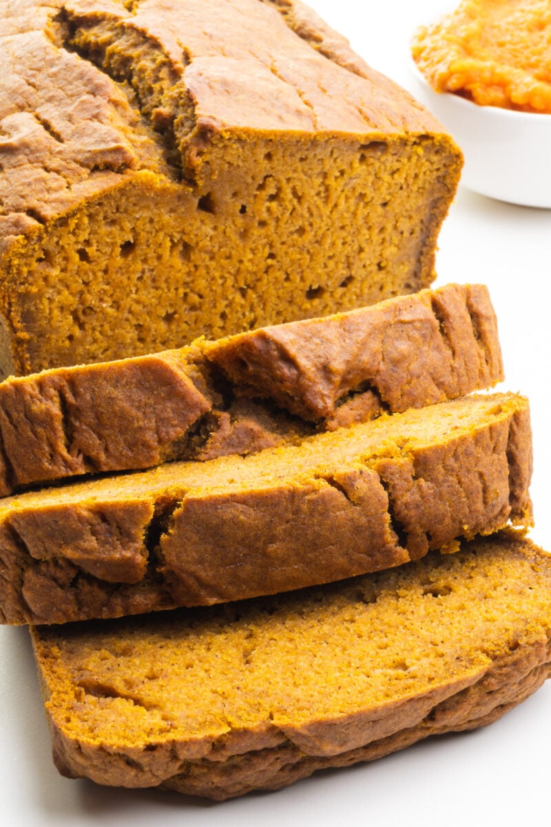 A loaf of vegan pumpkin bread has several slices cut. There's a bowl of Pumpkin Puree next to the loaf.