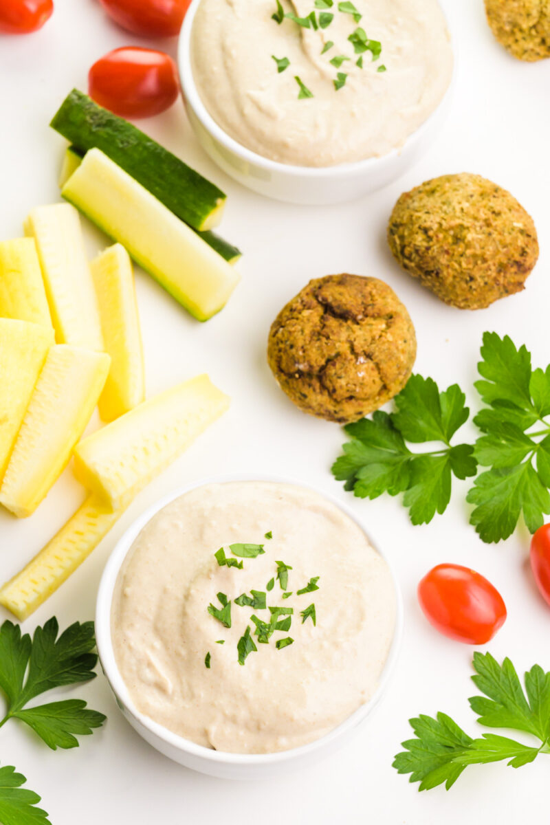 Looking down on two bowls of tahini sauce sitting next to cut up veggies, falafel, herbs, and tomatoes.
