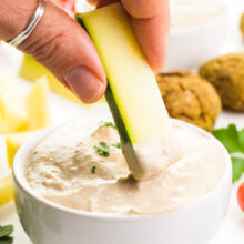 A hand holds a piece of zucchini and is dipping it in a creamy dip. Behind it are fresh herbs and other ingredients for dipping.