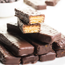 A stack of vegan Kit Kat Bars has two at the top that have been chopped in half, revealing the cookie underneath a chocolate coating. There are chocolate chips and more bars in the background.