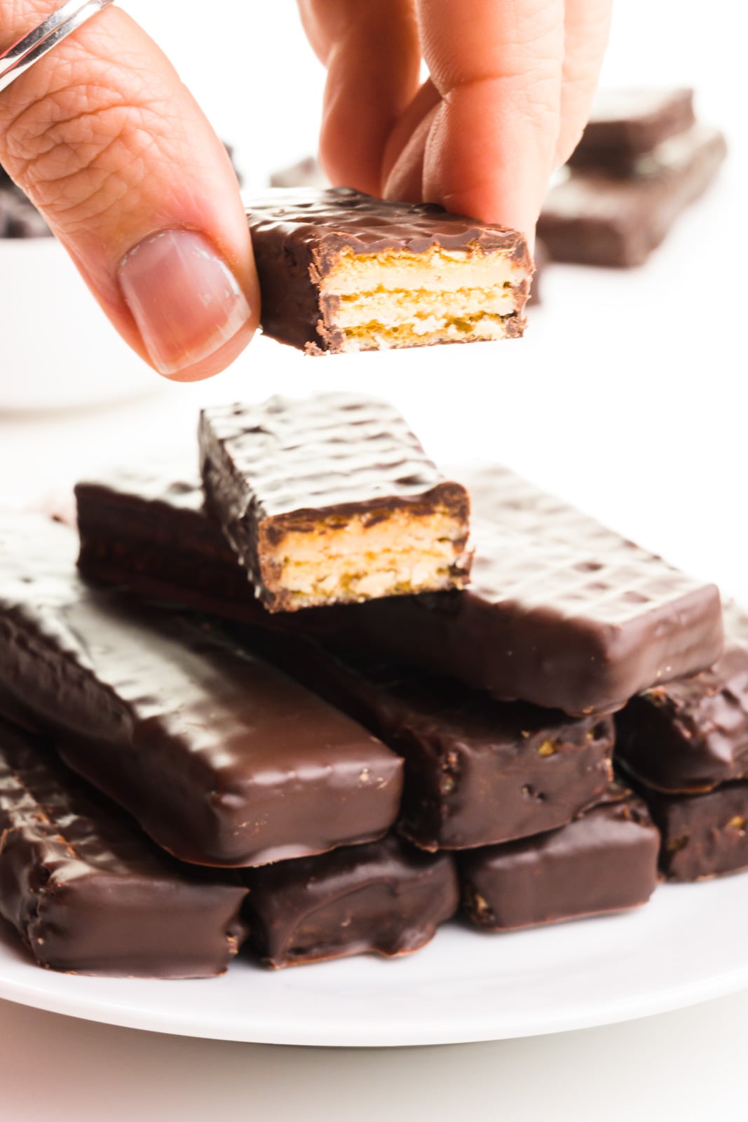 A hand holds a chocolate-coated cookie over a stack of more of them. Some are cut in half, showing the cookie inside the chocolate coating.