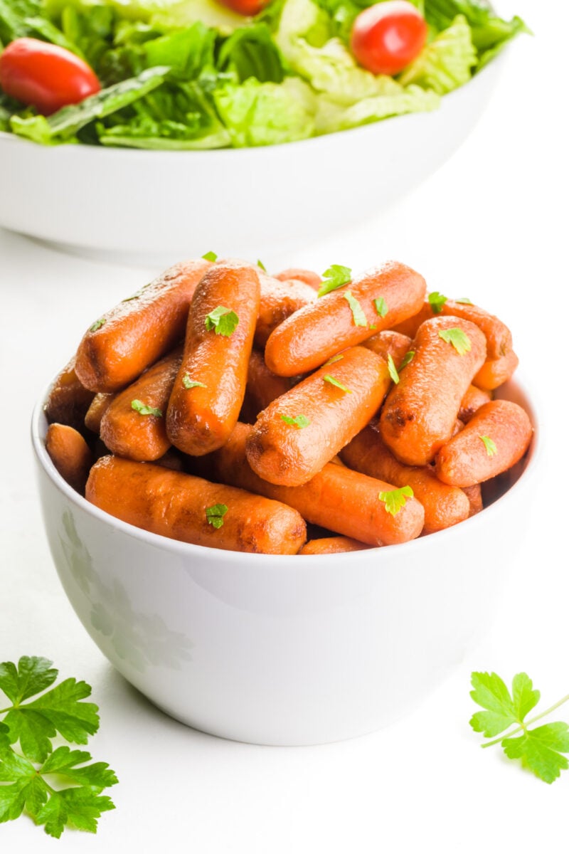 A bowl of roasted carrots has parsley on top and around it. There's a bowl of salad behind it.