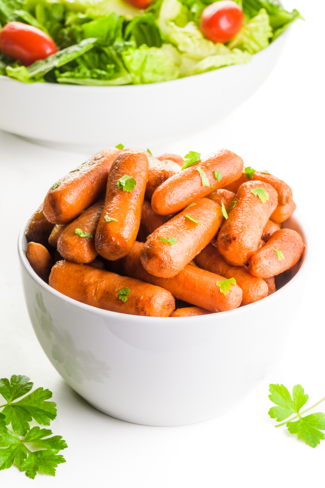 A bowl of roasted carrots has parsley on top and around it. There's a bowl of salad behind it.