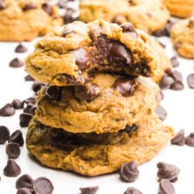 A stack of cookies shows the top one with a bite taken out. There are chocolate chips around it and more cookies in the background.