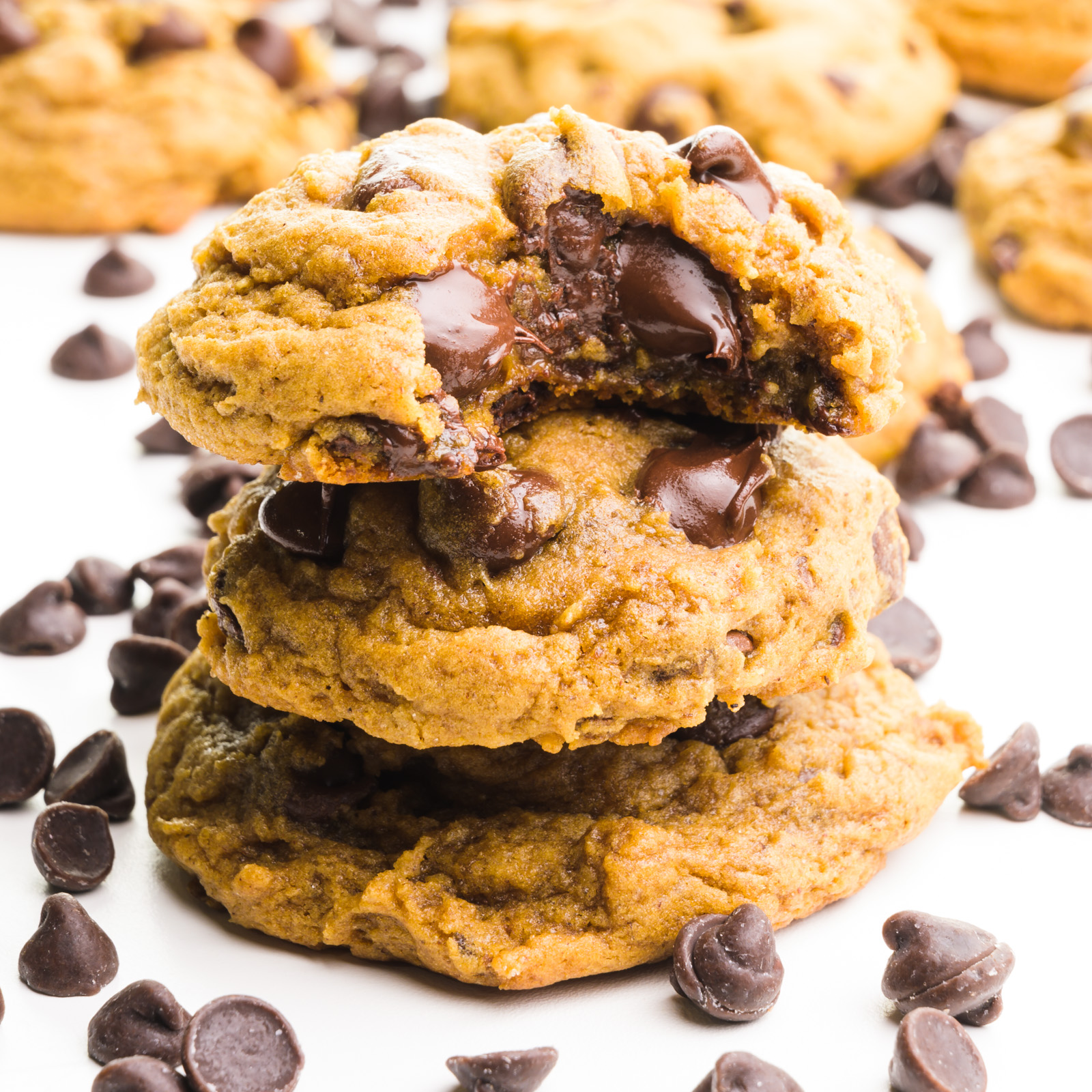 A stack of cookies shows the top one with a bite taken out. There are chocolate chips around it and more cookies in the background.