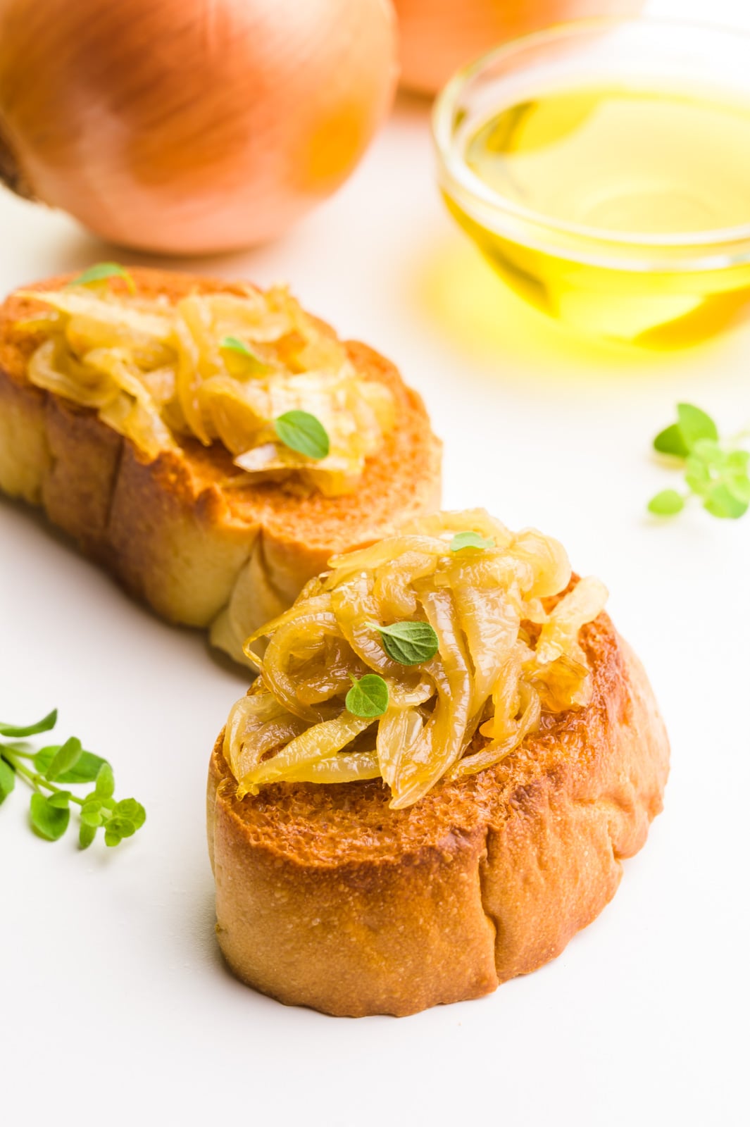Two slices of toasted baguettes are topped with onion jam and herbs. There are onions behind it and a bowl of olive oil.
