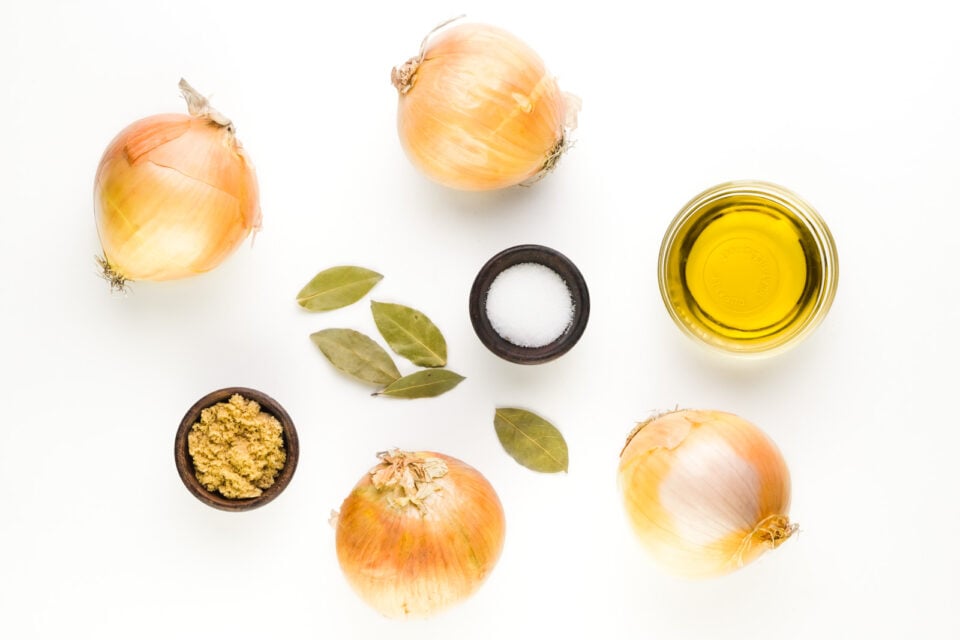 Ingredients are on a white counter, including bowls of olive oil and salt. There are several onions, bay leaves, and a bowl of brown sugar.