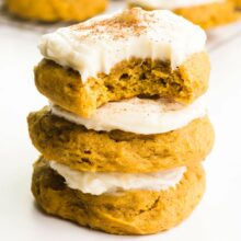 A stack of three cookies shows the top one with a bite taken out. Each cookie is frosted. There are more cookies in the background.