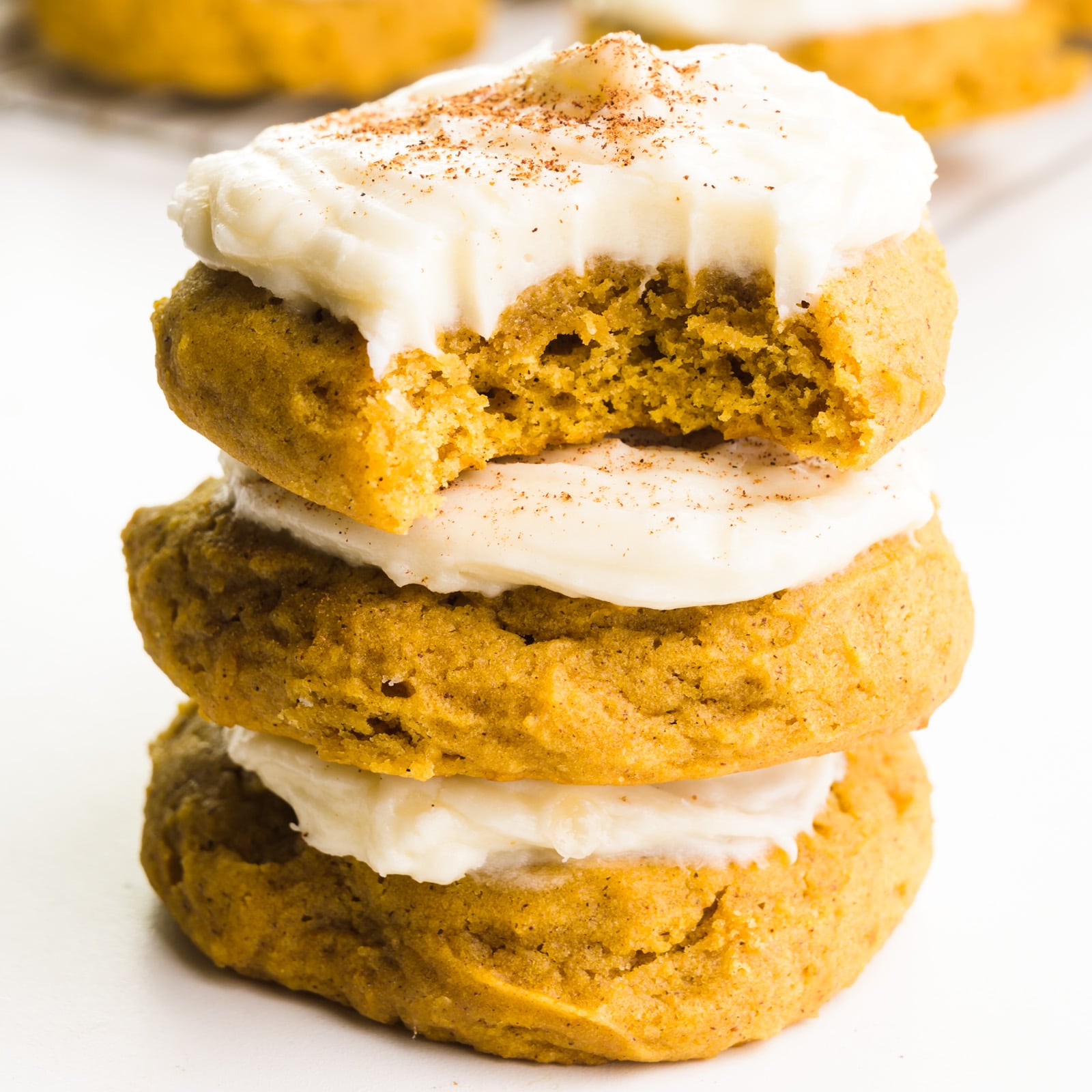 A stack of three cookies shows the top one with a bite taken out. Each cookie is frosted. There are more cookies in the background.