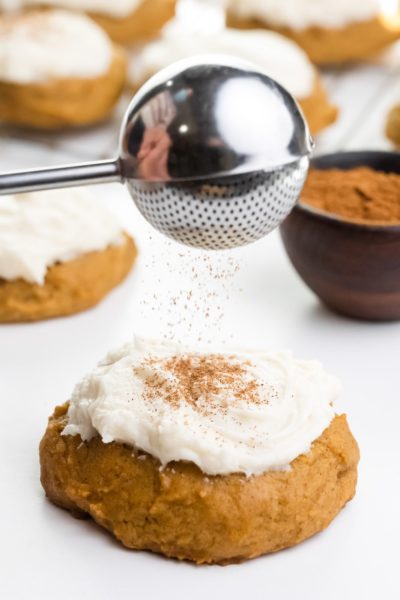 Spices are being sifted over a soft pumpkin cookie with frosting on top. There's a bowl of spices and more cookies behind it.