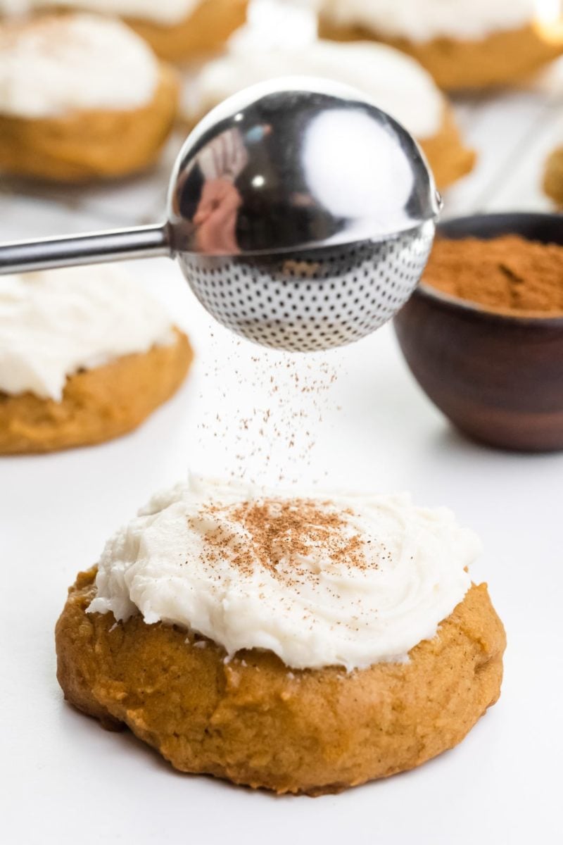 Spices are being sifted over a soft pumpkin cookie with frosting on top. There's a bowl of spices and more cookies behind it.
