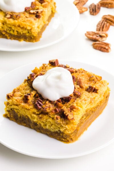 A pumpkin crisp bar sits on a white plate. It has a dollop of whipped cream on top. There's another bar on a plate and some nuts in the background.
