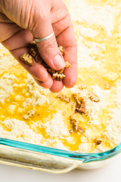 A hand distributes chopped pecans on top of cake batter in a pan.