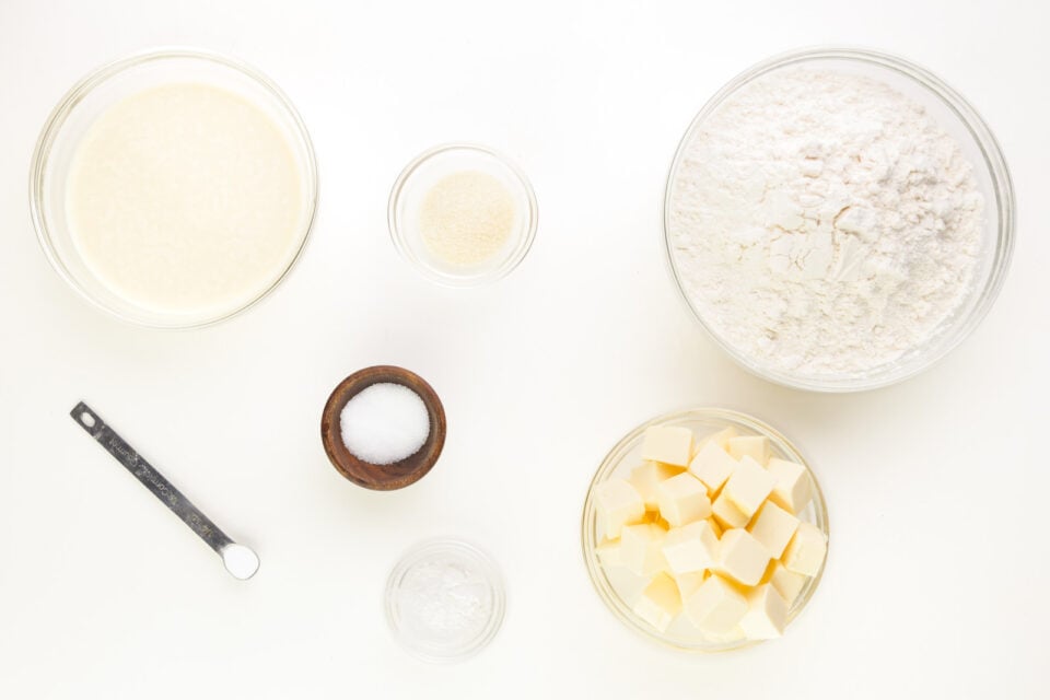 Bowls and measuring spoons are full of ingredients to make biscuits, like cubed butter, flour, baking powder, and more.
