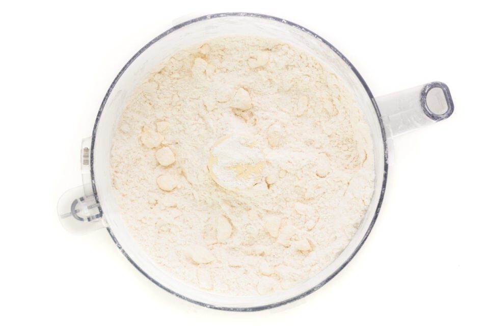 Looking down on a food processor bowl full of flour. There are little bits of butter distributed throughout.