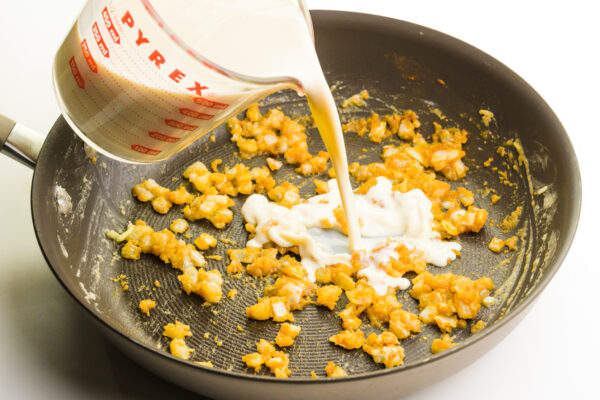 Vegan milk is being poured into a skillet with cooked onions and flour.