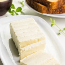 Vegan butter is on a plate. Several slices have been cut out. A piece of toast is in the background. A wooden bowl holds melted butter with herbs beside it.