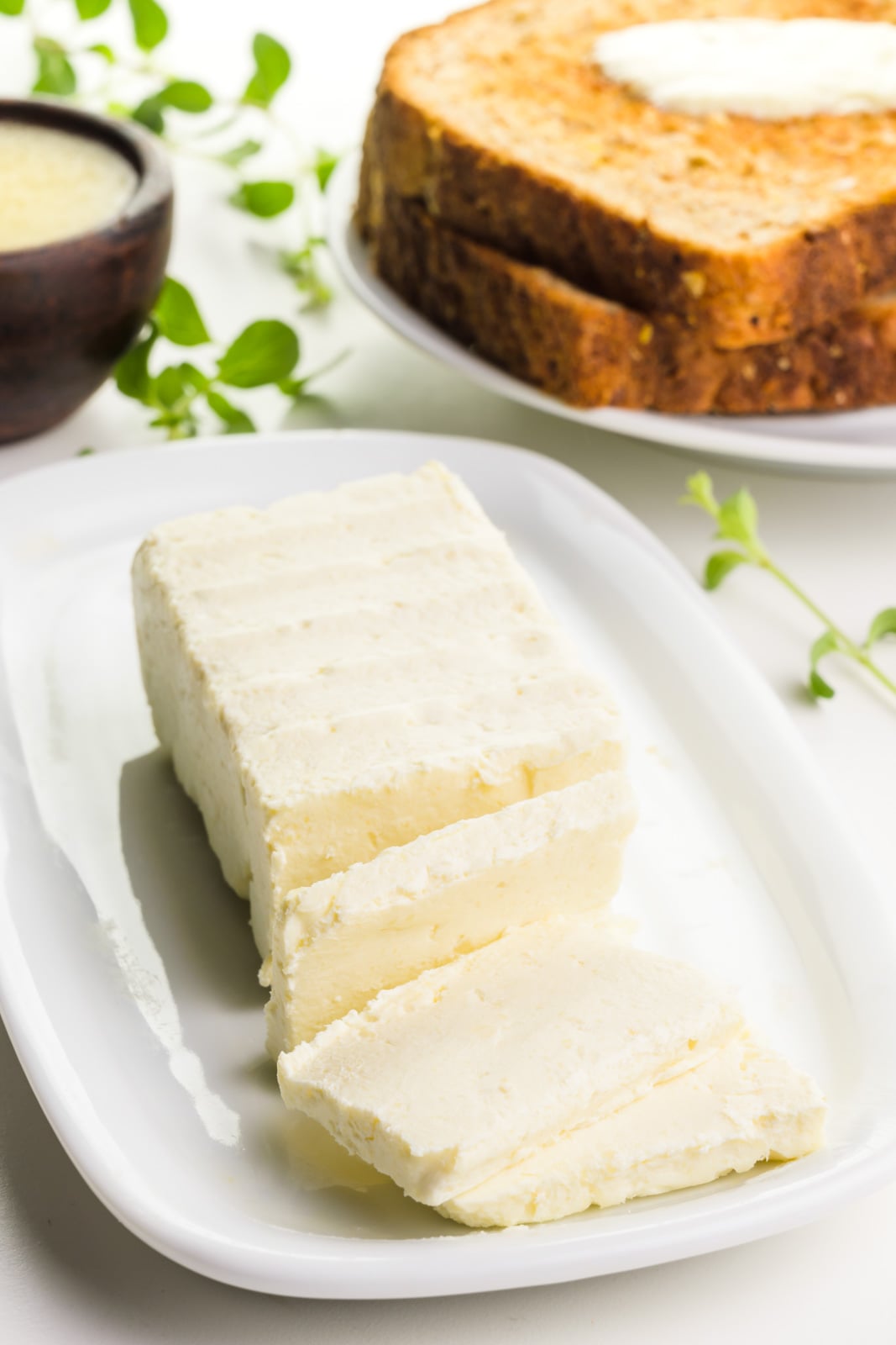 Vegan butter is on a plate. Several slices have been cut out. A piece of toast is in the background. A wooden bowl holds melted butter with herbs beside it.